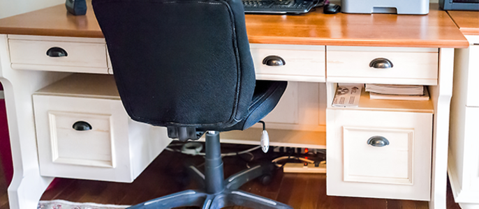 Ein Heimbüro mit einem Holzschreibtisch, einem schwarzen Drehstuhl, einem Computermonitor und verschiedenen Bürogeräten.