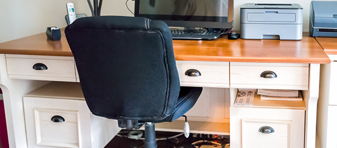 Ein Heimbüro mit einem Holzschreibtisch, einem schwarzen Drehstuhl, einem Computermonitor und verschiedenen Bürogeräten.