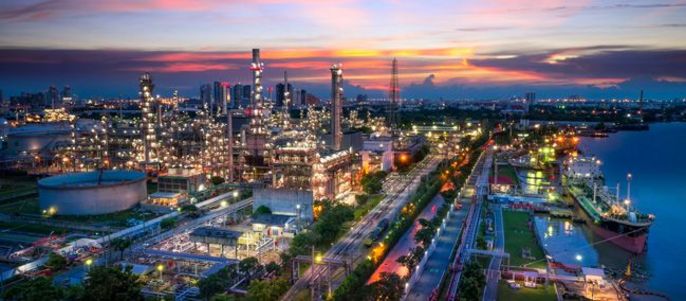 Ein Industriegebiet bei Sonnenuntergang mit einer Raffinerie mit mehreren Schornsteinen, Lagertanks und Straßen sowie einem Fluss, an dem ein Schiff angedockt ist.