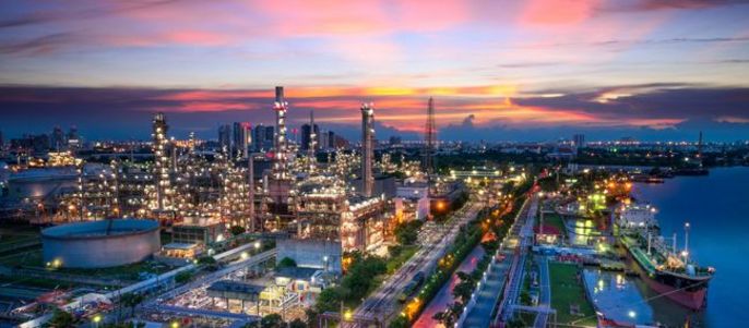 Ein Industriegebiet bei Sonnenuntergang mit einer Raffinerie mit mehreren Schornsteinen, Lagertanks und Straßen sowie einem Fluss, an dem ein Schiff angedockt ist.