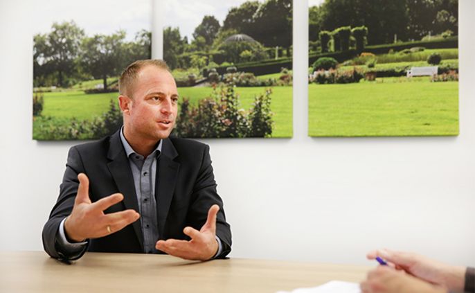 Ein Mann im Anzug, der mit seinen Händen gestikuliert, in einem Raum mit Bildern eines Gartens an der Wand.