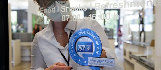 Eine Frau in weißem Hemd und Maske bringt einen blauen Aufkleber an einer Glastür an, der darauf hinweist, dass der Raum gegen Corona abgesichert ist.