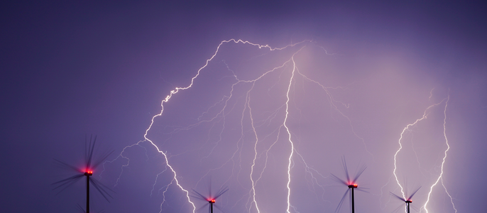 Ein dramatischer Nachthimmel mit zahlreichen Blitzen, während sich mehrere Windturbinen als Silhouette vom Hintergrund abheben.