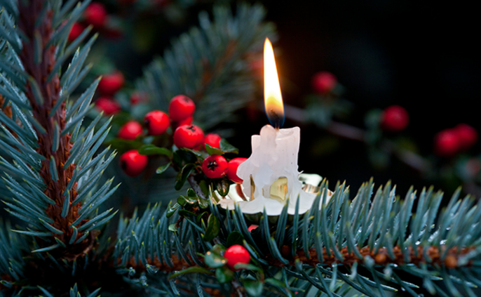 Eine brennende Kerze auf einem Zweig mit roten Beeren, umgeben von grünen Nadeln.