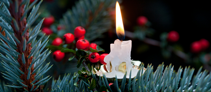 Eine brennende Kerze auf einem Zweig mit roten Beeren, umgeben von grünen Nadeln.