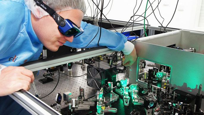 Eine Person in einem blauen Laborkittel, die eine Schutzbrille trägt und einen komplexen Apparat mit grün leuchtenden Bauteilen genau untersucht.