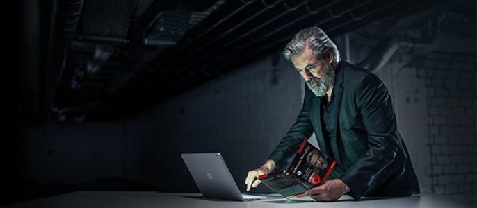 Ein älterer Mann mit Bart, der eine Brille trägt und in einem schwach beleuchteten Raum mit freiliegenden Rohren an der Decke an einem Laptop arbeitet.