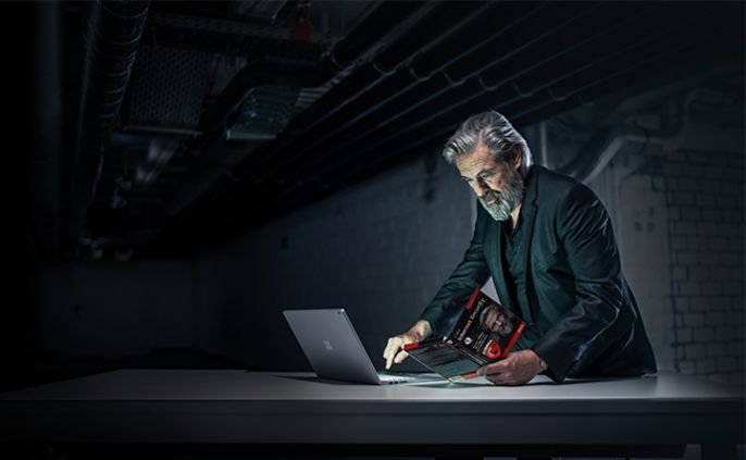 Ein älterer Mann mit Bart, der eine Brille trägt und in einem schwach beleuchteten Raum mit freiliegenden Rohren an der Decke an einem Laptop arbeitet.