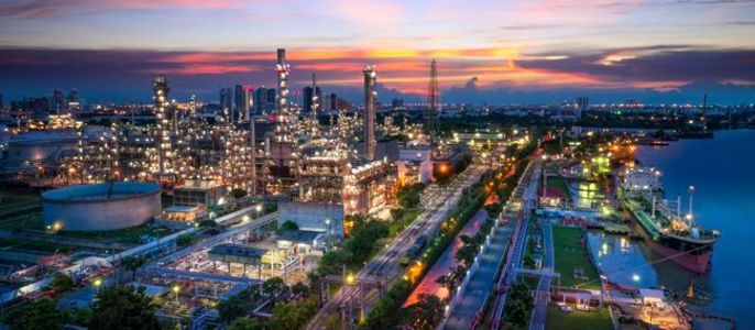 Ein Industriegebiet bei Sonnenuntergang mit einer Raffinerie mit mehreren Schornsteinen, Lagertanks und Straßen sowie einem Fluss, an dem ein Schiff angedockt ist.