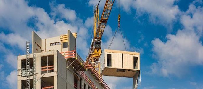 Eine Baustelle, auf der ein Kran einen Betonfertigteil eines Gebäudes anhebt.