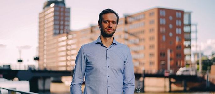 Ein Mann in einem blauen Hemd, der an einem Fluss steht, mit Gebäuden im Hintergrund.