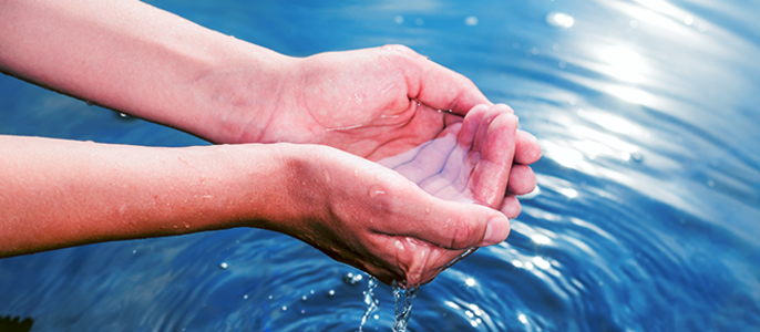 Die Hände einer Person halten Wasser, welches heruntertropft und Wellen in einem Bach darunter bilden.