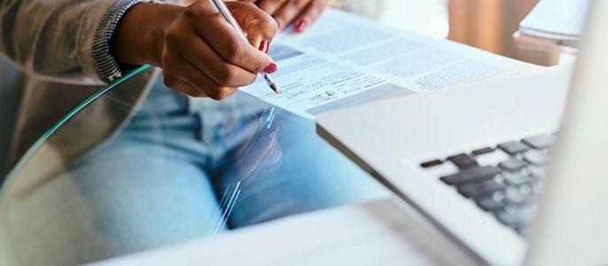 Die Hände einer Person, die auf einem Papier schreibt, mit einem Laptop und einigen Papieren in der Nähe.