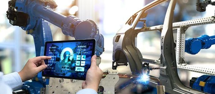 Eine Person bedient ein Tablet, auf dem ein blauer Roboterarm in einer Fabrikumgebung abgebildet ist, der einen Fahrzeugrahmen schweißt.