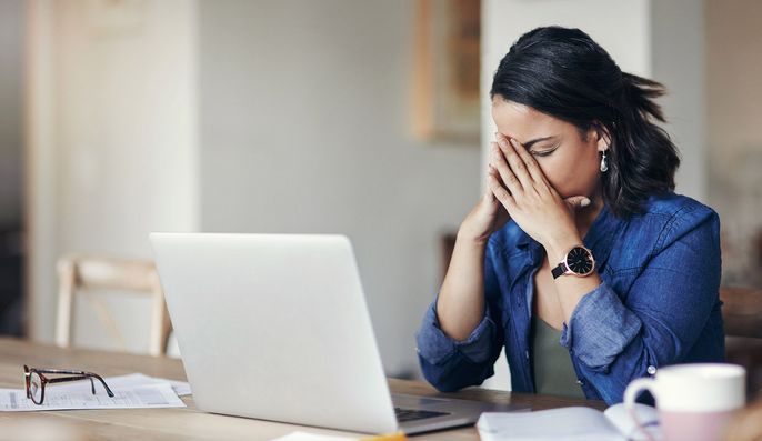 Eine Frau mit dunklem Haar und blauem Hemd sitzt an einem Tisch mit einem Laptop, einer Brille und einer Tasse. Ihre Hände bedecken ihr Gesicht in einer Geste der Verzweiflung.