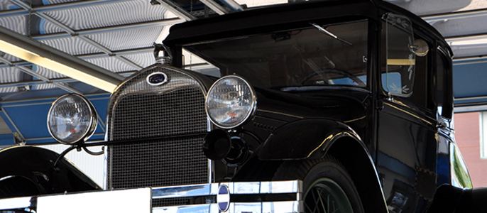 Ein schwarzer Oldtimer mit runden Scheinwerfern, einem auffälligen Kühler und einem Nummernschildhalter, der unter einer Metallstruktur geparkt ist.