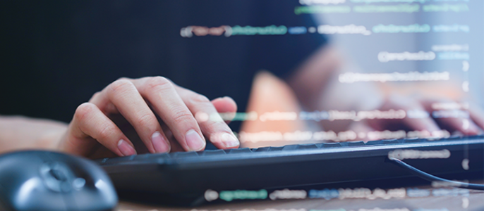 Eine Person, die auf einer Tastatur tippt, mit einer Computermaus neben sich, umgeben von Codezeilen.