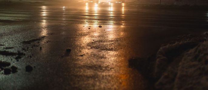 Eine nasse Straße bei Nacht, die Scheinwerfer der Fahrzeuge reflektieren auf der nassen Oberfläche, und in der Ferne läuft eine Person.