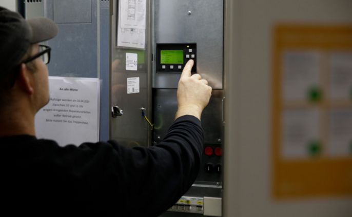 Ein Mann mit schwarzem Hemd und Brille zeigt auf eine digitale Anzeige an einer Maschine. An der Maschine ist ein Papier mit einem Text in deutscher Sprache befestigt.