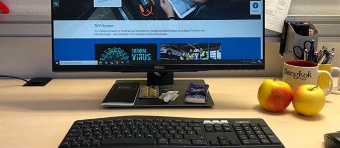 Ein Arbeitsbereich mit einem Computerbildschirm, auf dem eine Website angezeigt wird, einer Tastatur, einer Maus, einer Tasse mit einem Logo und zwei Äpfeln auf dem Schreibtisch.