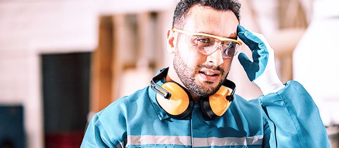 Ein Mann in einer blauen Arbeitsuniform, mit Schutzbrille und Handschuhen, rückt seine Brille zurecht und schaut in die Kamera.