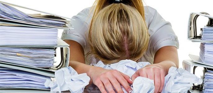 Eine Frau mit blondem Haar, die ein weißes Hemd trägt, umgeben von Papierstapeln und zerknitterten Papieren auf einem Schreibtisch.