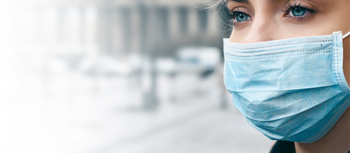 Nahaufnahme eines Frauengesichts mit einer blauen chirurgischen Maske vor einem verschwommenen Hintergrund einer Stadtlandschaft.