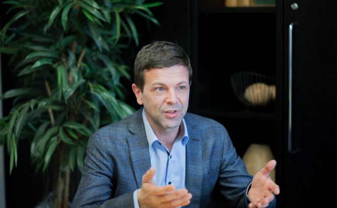 Ein Mann in einem blauen Blazer, der mit geöffneten Händen spricht, in einem Innenraum mit einer Pflanze und dekorativen Gegenständen im Hintergrund.