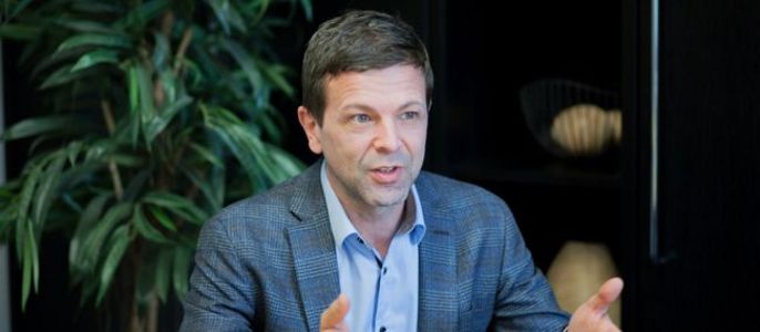 Ein Mann in einem blauen Blazer, der mit geöffneten Händen spricht, in einem Innenraum mit einer Pflanze und dekorativen Gegenständen im Hintergrund.