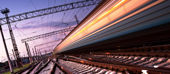 Ein schnell fahrender Zug auf Gleisen, mit Stromleitungen über dem Kopf und einem violetten Himmel im Hintergrund.
