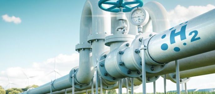 Ein großes Rohrsystem mit der Aufschrift "H2" und einem Messgerät auf einem offenen Feld mit Windkraftanlagen im Hintergrund.