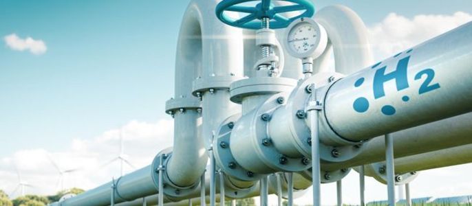 Ein großes Rohrsystem mit der Aufschrift "H2" und einem Messgerät auf einem offenen Feld mit Windkraftanlagen im Hintergrund.