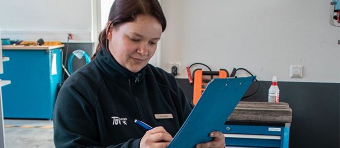 Eine Frau in einer Werkstatt, die eine schwarze Jacke mit einem Logo trägt und auf einem blauen Klemmbrett schreibt.
