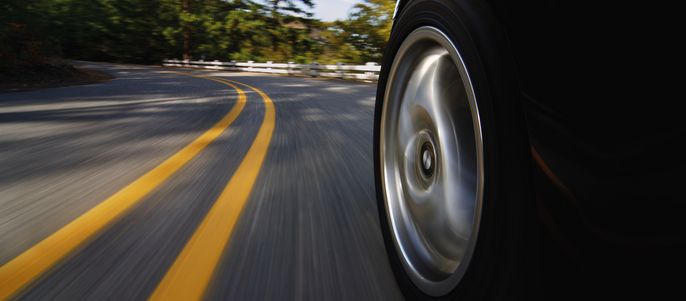 Ein Autoreifen in Bewegung auf einer kurvenreichen Straße, umgeben von Bäumen und Hügeln.