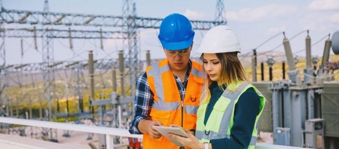 Zwei Arbeiter, ein Mann und eine Frau, die Sicherheitswesten und Helme tragen, untersuchen ein Tablet-Gerät in einer Außenumgebung mit elektrischen Geräten im Hintergrund.