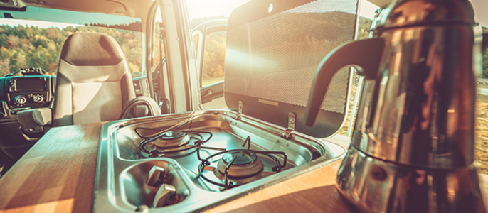 Das Innere eines Fahrzeugs mit einem Herd und einer Kaffeemaschine vor einer malerischen Kulisse aus Bäumen und einem sonnenbeschienenen Himmel.