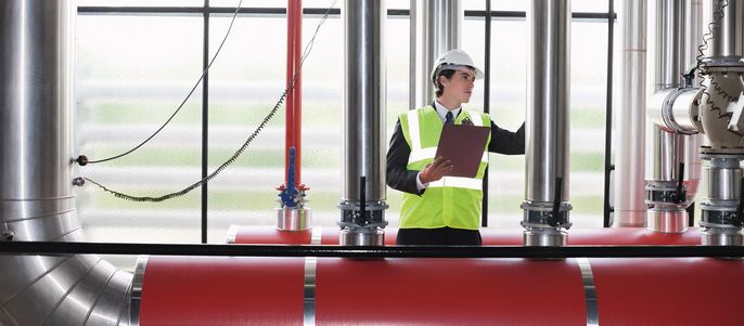 Eine Industrieumgebung mit einem Mann mit Schutzhelm und reflektierender Weste, der große Rohre und Maschinen inspiziert.