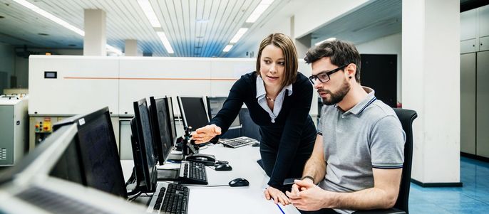 Zwei Personen in einer modernen Büroumgebung, wobei eine Person auf einen Computerbildschirm zeigt, während die andere zusieht.