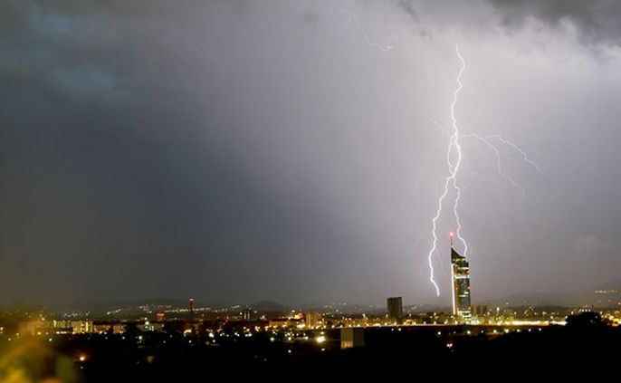 Eine nächtliche Stadtlandschaft mit einem markanten Blitz, der in ein hohes Gebäude einschlägt.