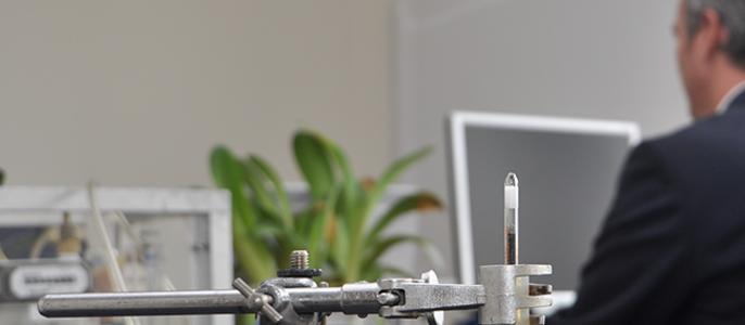 Eine Laboreinrichtung mit einem Glasrohr, das an einem Metallständer befestigt ist, und einem Mann im Anzug, der im Hintergrund an einem Computer arbeitet.