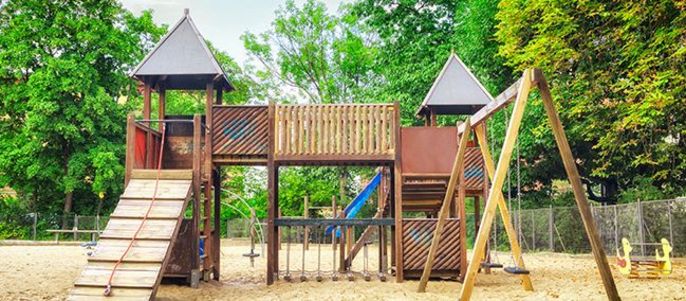 Ein hölzerner Spielplatz mit Rutsche, Klettergerüst und Schaukel vor der Kulisse grüner Bäume und eines klaren Himmels.
