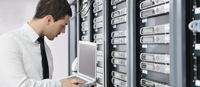 Ein Mann in weißem Hemd und Krawatte, der vor einer Reihe von Servern an einem Laptop arbeitet.