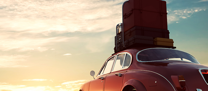 Ein roter Oldtimer, der bei Sonnenuntergang auf einem freien Feld geparkt ist und auf dessen Dach ein Stapel Koffer liegt.