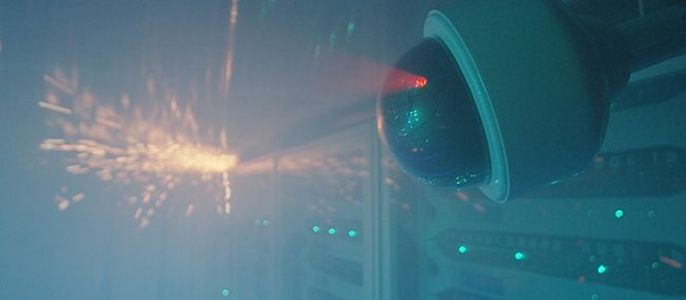 Eine Überwachungskamera mit einem roten Laserstrahl vor der Kulisse eines Serverraums mit mehreren Lichtern und Dunst.