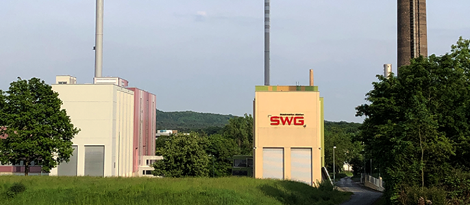 Zwei Industriegebäude mit hohen Schornsteinen, eines davon mit dem Logo "SWG", umgeben von Grünflächen und einem Weg.