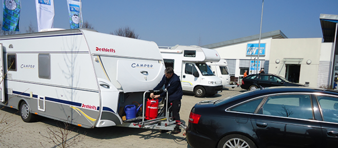 Ein weißer Wohnwagen. Ein Mann ist in der Nähe des Wohnwagens zu sehen, und im Hintergrund sind weitere Fahrzeuge und ein Geschäft zu sehen.