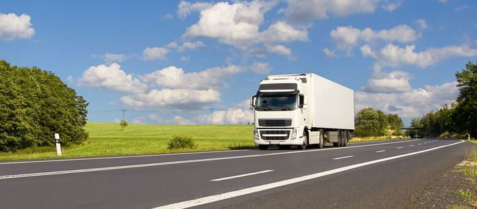 Ein weißer Lastwagen fährt auf einer Straße, die von grünen Feldern und Bäumen umgeben ist, unter einem blauen Himmel mit weißen Schäfchenwolken.