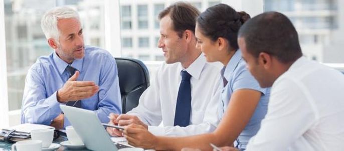 Eine Gruppe von vier Fachleuten in einem modernen Büro, die an einem Tisch mit einem Laptop und einer Tasse Kaffee diskutieren.