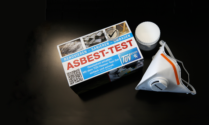 Eine Schachtel mit der Aufschrift 'RENOVIEREN - SANIEREN - UMBAUEN' und dem Titel 'ASBEST-TEST'. Daneben liegt eine weiße FFP-2 Maske.