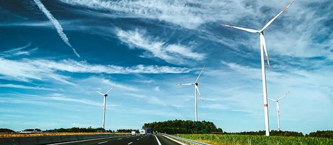 Ein strahlend blauer Himmel mit weißen Wolken, eine mehrspurige Autobahn und drei hohe Windräder am Straßenrand.